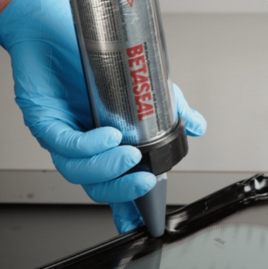 A person in blue gloves uses a spray gun to apply Betaseal sealant on a glass window at Epic Windscreens.