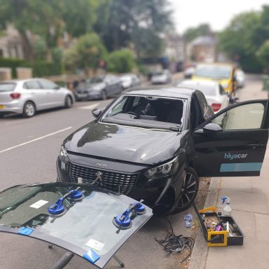 A Peugeot 208 is being repaired on the roadside, getting a windscreen replacement for a fleet service.