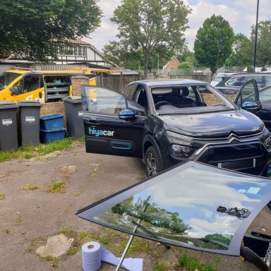 A fleet car with a broken windshield parked in a driveway, surrounded by vehicles and an Epic Windscreens van under clear blue skies.